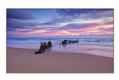 Beach scene with a shipwreck at sunset, featuring a colorful sky.