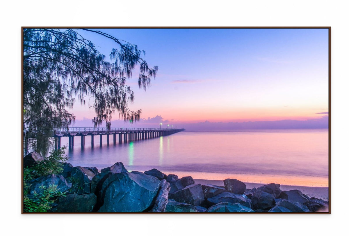Pier extending into the water with a colorful sunset sky, framed by a tree on the left. wall art print