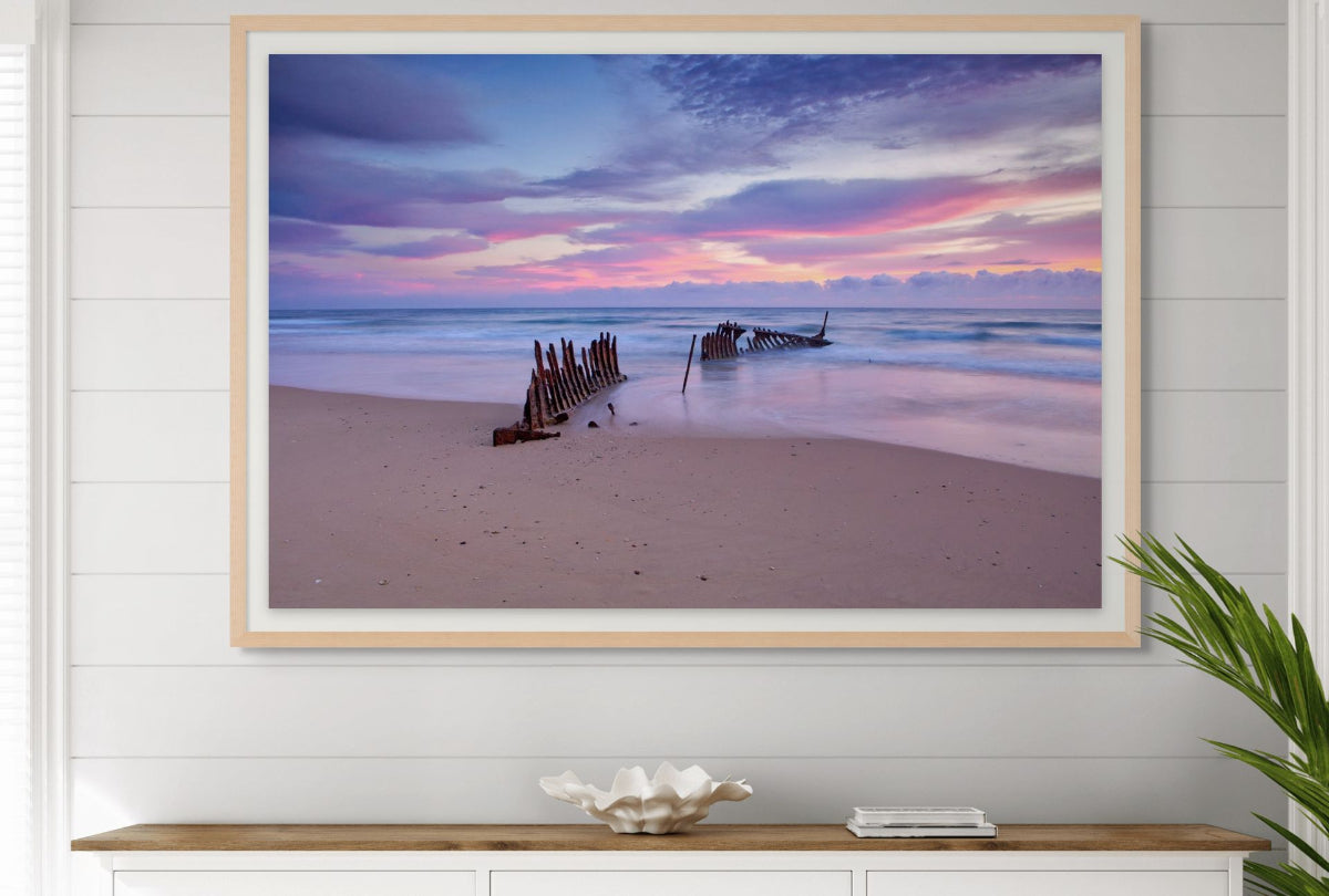 Framed beachscape with sunset over water and shipwreck on sand, displayed on a wall.