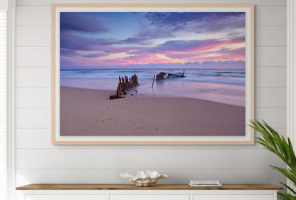 Framed beachscape with sunset over water and shipwreck on sand, displayed on a wall.