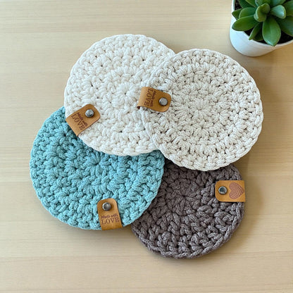 Four handmade crocheted coasters in natural, beige, blue and gray with leather tags.