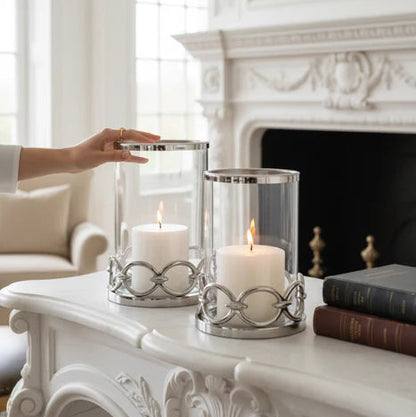 Decorative candles in silver hurricane holders on a white mantelpiece with a fireplace in the background.