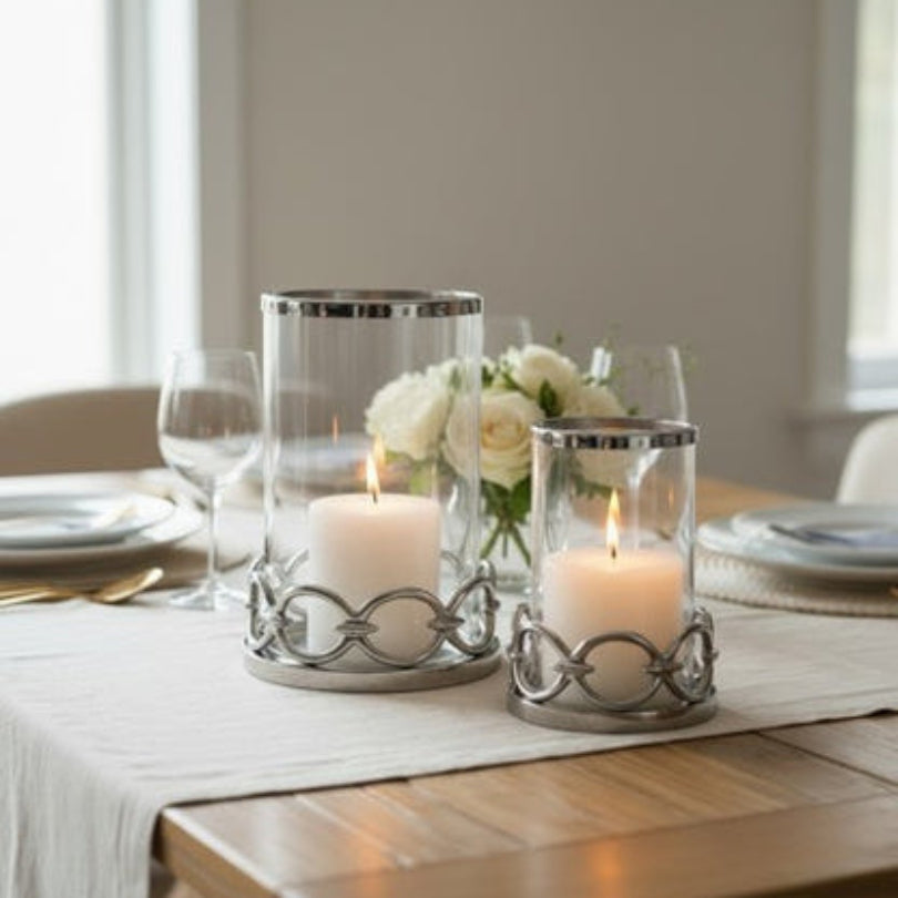 Decorative candles in silver and glass hurricane holders on a table with white flowers and plates.